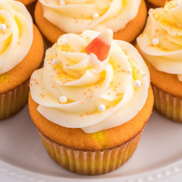 Square close-up of Candy Corn Cupcake topped with swirled white frosting, candy corn, and sugar pearls on a white plate.