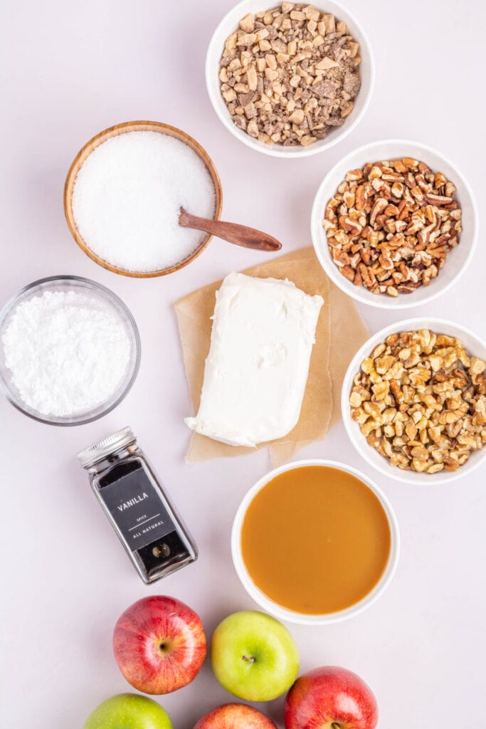 Overhead view of ingredients for caramel apple dip including cream cheese, caramel sauce, nuts, toffee bits, powdered sugar, vanilla, and apples.