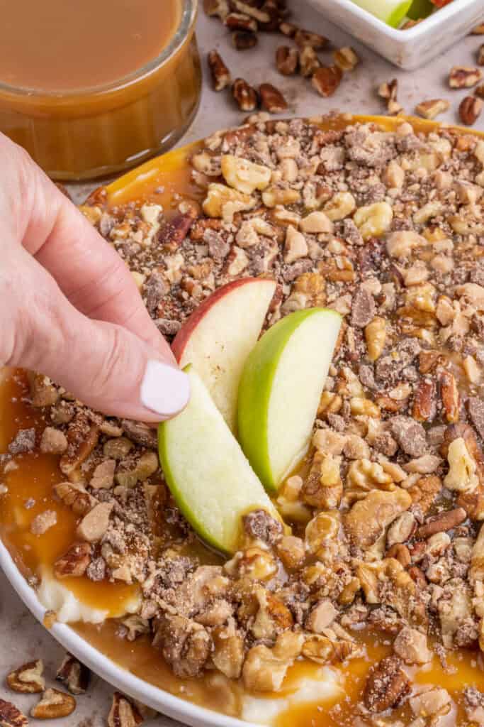 Hand dipping apple slices into caramel apple dip topped with nuts and toffee pieces.
