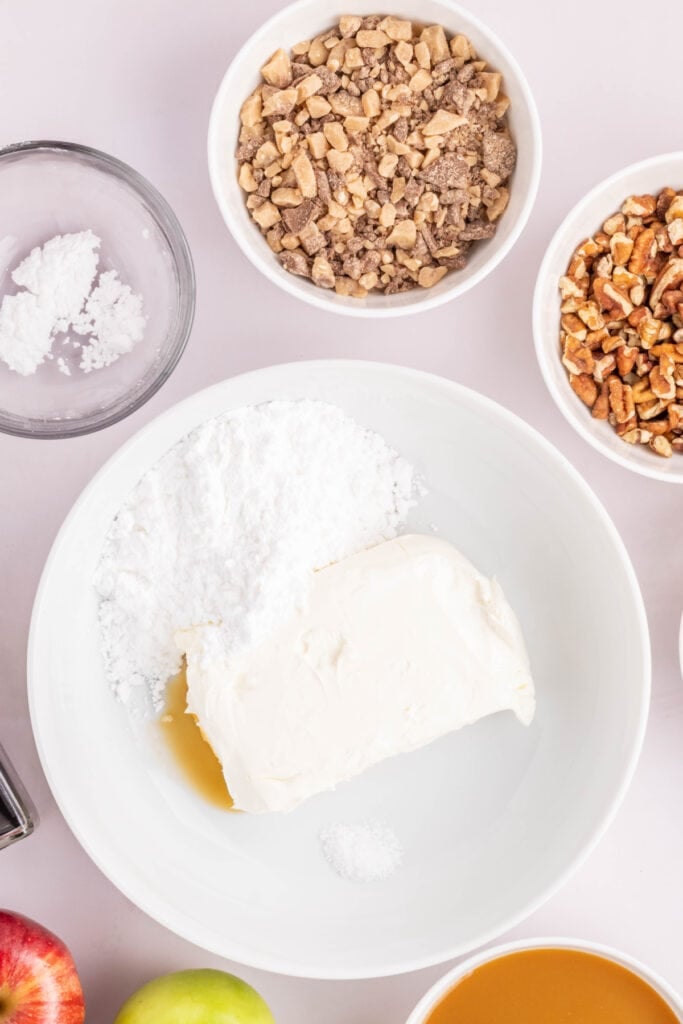 Mixing bowl with cream cheese, powdered sugar, vanilla, and salt surrounded by bowls of nuts and toffee bits.