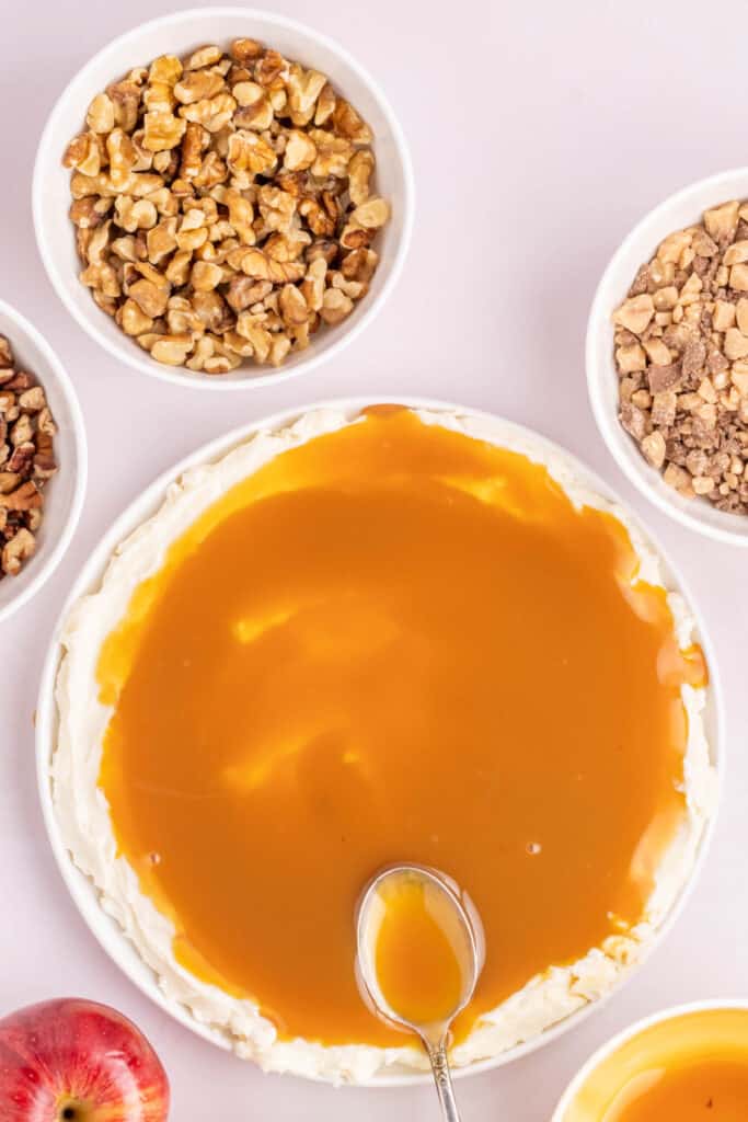 Caramel sauce being spooned over the cream cheese layer in a round dish.