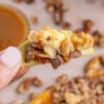 Square image of caramel apple dip topped with nuts and toffee bits on top of an apple slice held by a hand