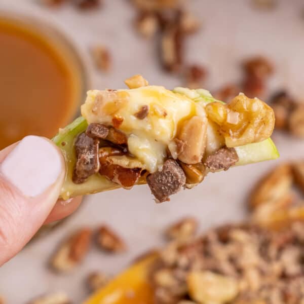 Square image of caramel apple dip topped with nuts and toffee bits on top of an apple slice held by a hand