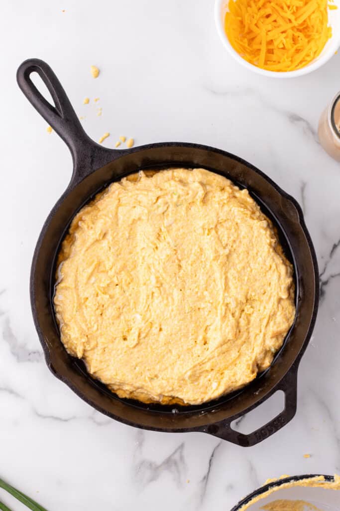 Unbaked Cheddar Onion Cornbread batter spread evenly in a hot cast iron skillet, ready for baking.