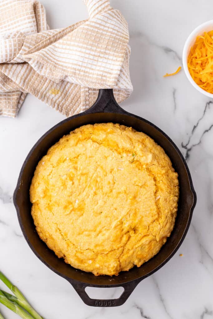 Freshly baked Cheddar Onion Cornbread in a cast iron skillet with a golden top, cooling on a marble counter.