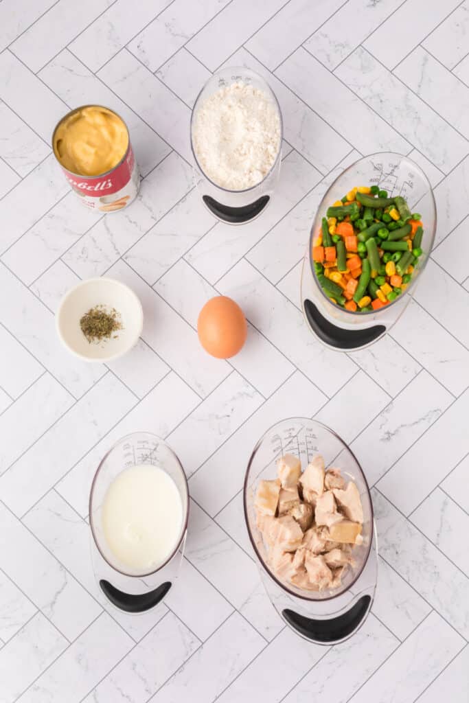 Overhead view of Chicken Biscuit Pot Pie ingredients measured out, including cream of chicken soup, biscuit mix, milk, egg, chicken, vegetables, and thyme on a white herringbone surface.