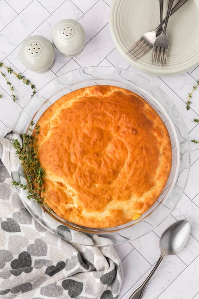 Golden brown baked Chicken Biscuit Pot Pie fresh from the oven with sprigs of thyme and serving utensils beside it.