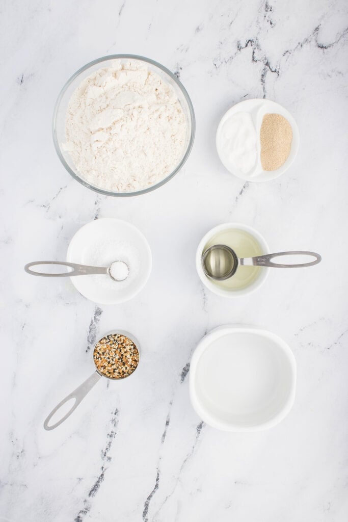 Overhead view of measured ingredients for everything bagel no knead bread arranged on a marble surface.