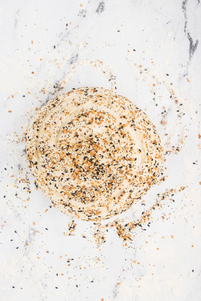 Overhead shot of unbaked everything bagel dough sprinkled with seasoning on a lightly floured surface.