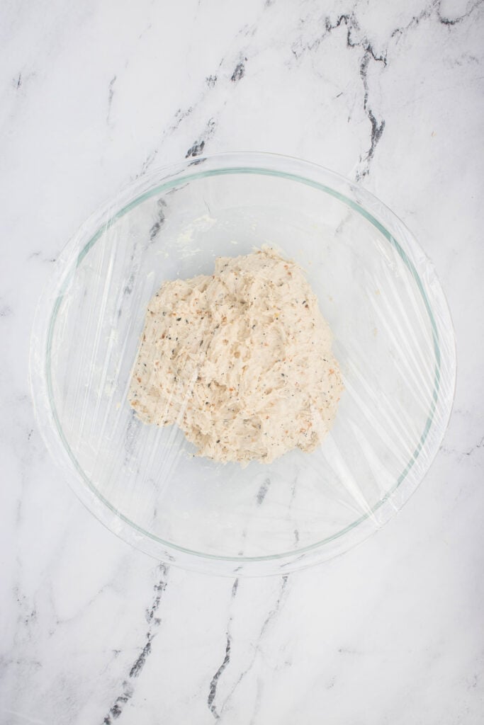 Bowl of everything bagel dough covered with plastic wrap to rest and rise.