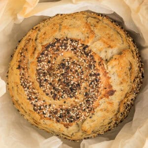 Overhead close-up of golden baked everything bagel no knead bread topped with sesame, poppy, and garlic seasoning in parchment paper.