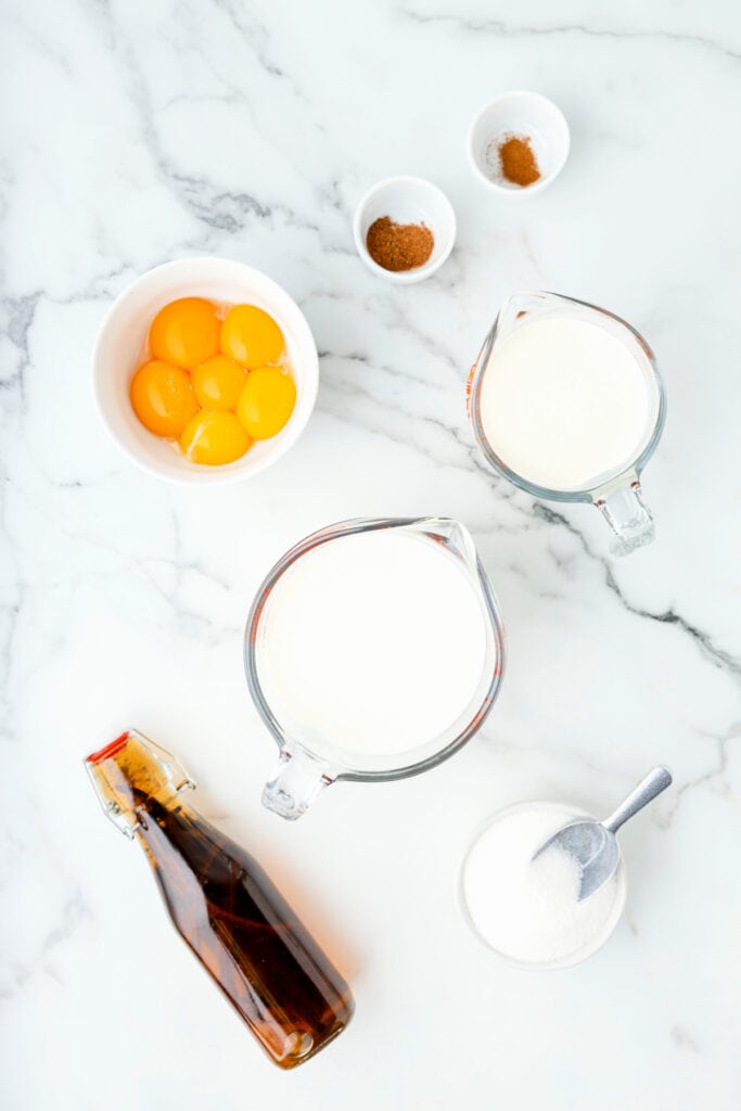 Overhead view of eggnog ingredients including egg yolks in a bowl, sugar, milk, cream, spices, and vanilla on a marble counter.