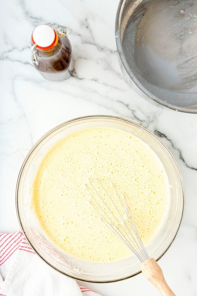 Whisk resting in a bowl of tempered egg and milk mixture, smooth and frothy on top.