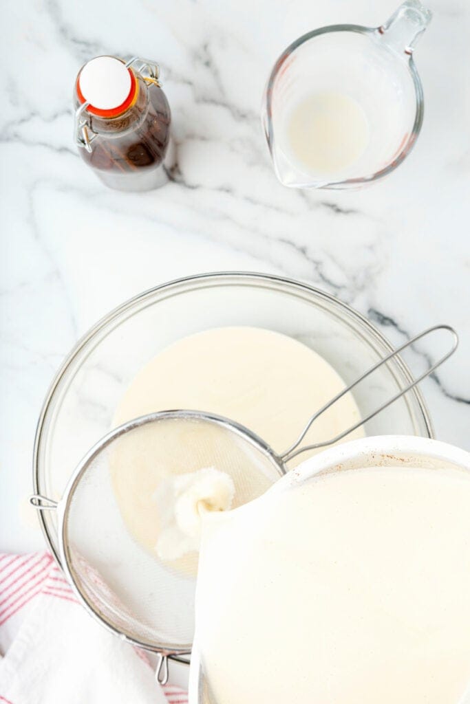 Eggnog being poured through a fine mesh strainer into a clean glass bowl.