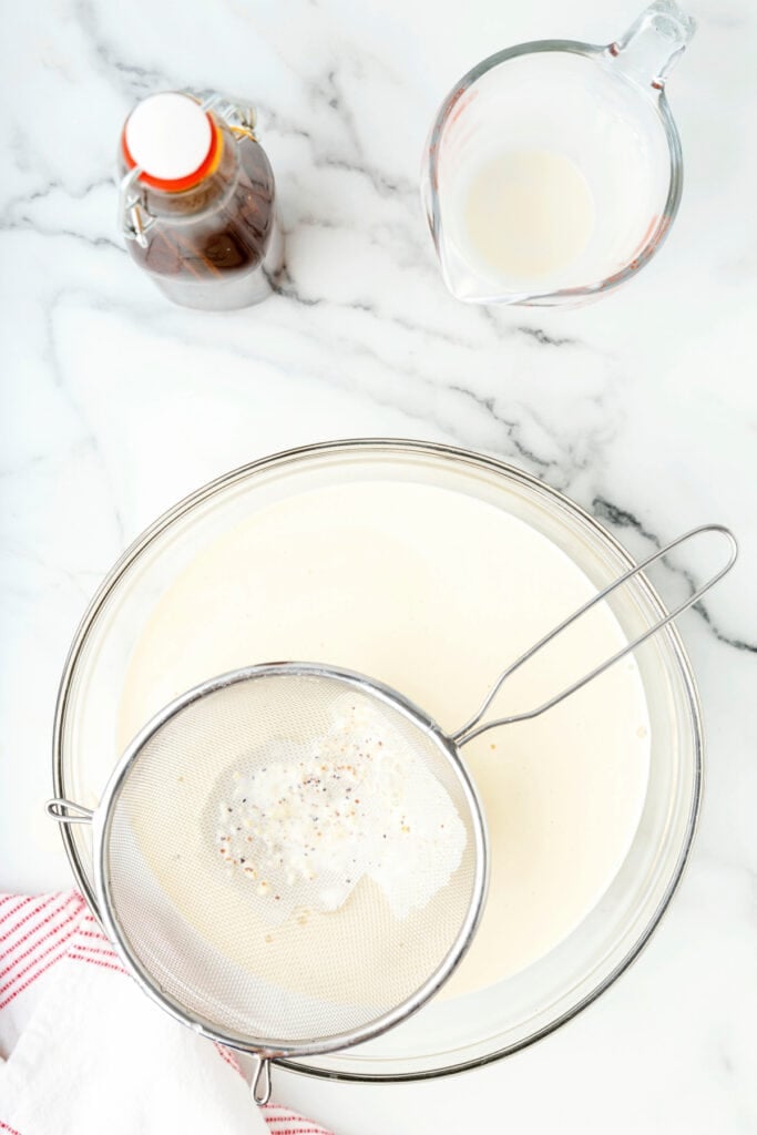 Mesh strainer catching spices and solids as eggnog passes through into a glass bowl.