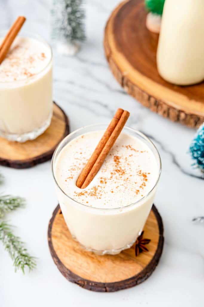 Glass of homemade eggnog on a wood coaster, topped with ground nutmeg and a cinnamon stick, with holiday décor in background.
