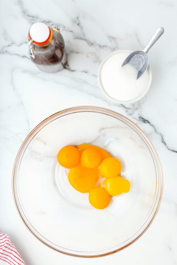 Glass bowl with raw egg yolks ready to be mixed, with sugar and vanilla nearby on the counter.