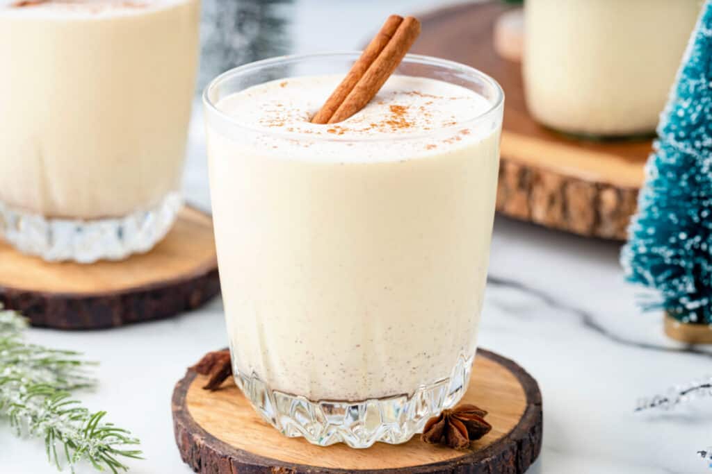 Glass of homemade eggnog topped with ground nutmeg and a cinnamon stick, served on a wood coaster with festive holiday décor.