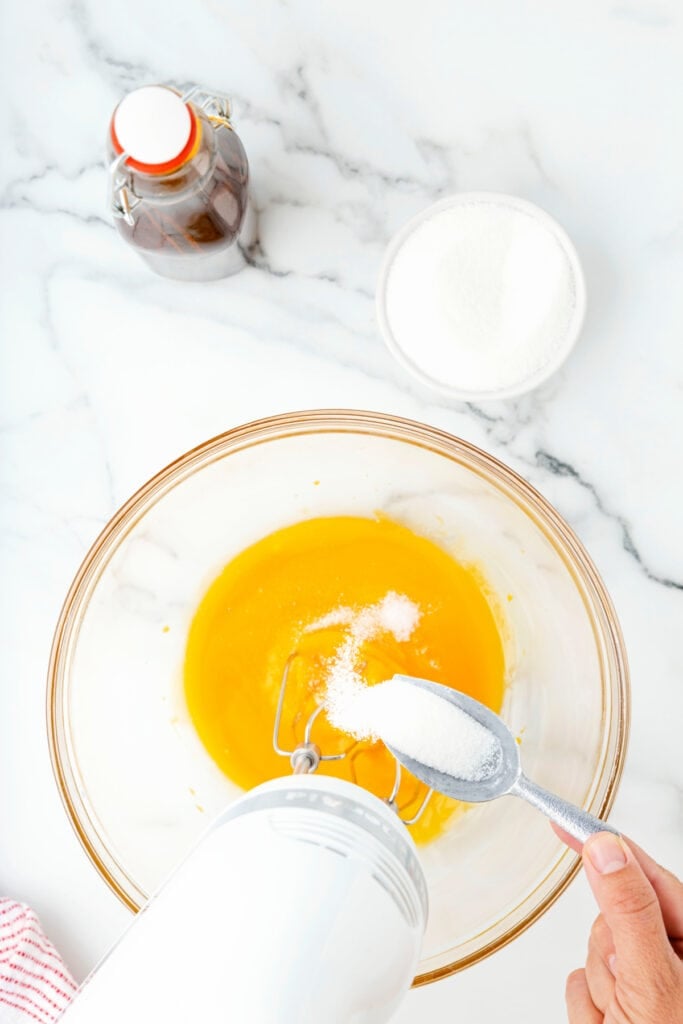 Sugar being spooned into egg yolks in a glass mixing bowl with an electric mixer.