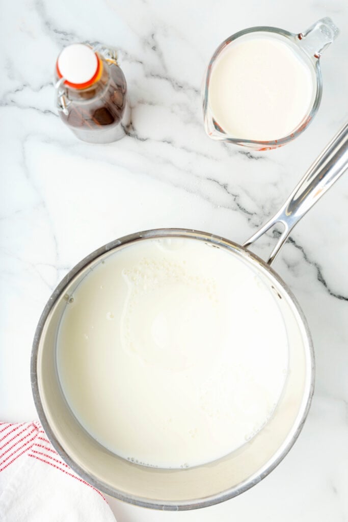 Saucepan filled with milk heating on the stove, with vanilla and cream nearby.