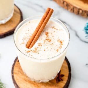 Overhead view of a glass of eggnog garnished with a cinnamon stick and nutmeg, styled with wood coaster and holiday greenery.