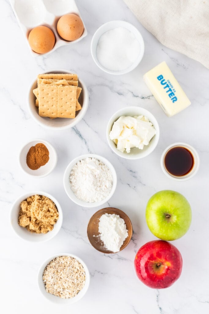 Overhead shot of ingredients for Mini Apple Crumble Cheesecakes including apples, cream cheese, eggs, graham crackers, oats, sugar, butter, and cinnamon on a marble surface.
