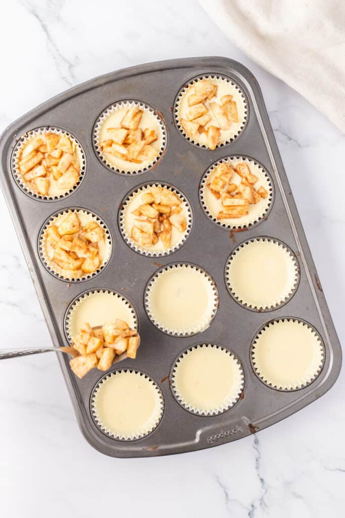 Muffin pan filled with mini cheesecakes being topped with spoonfuls of cinnamon-spiced apple mixture.