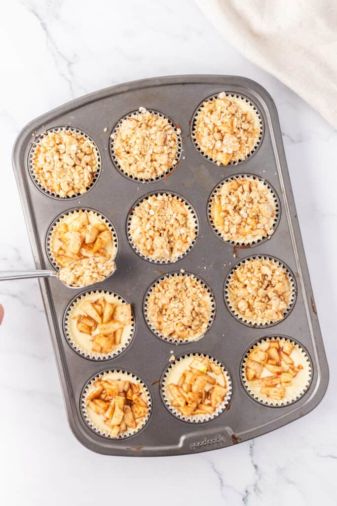 Muffin pan with mini apple cheesecakes being topped with oat crumble before baking.