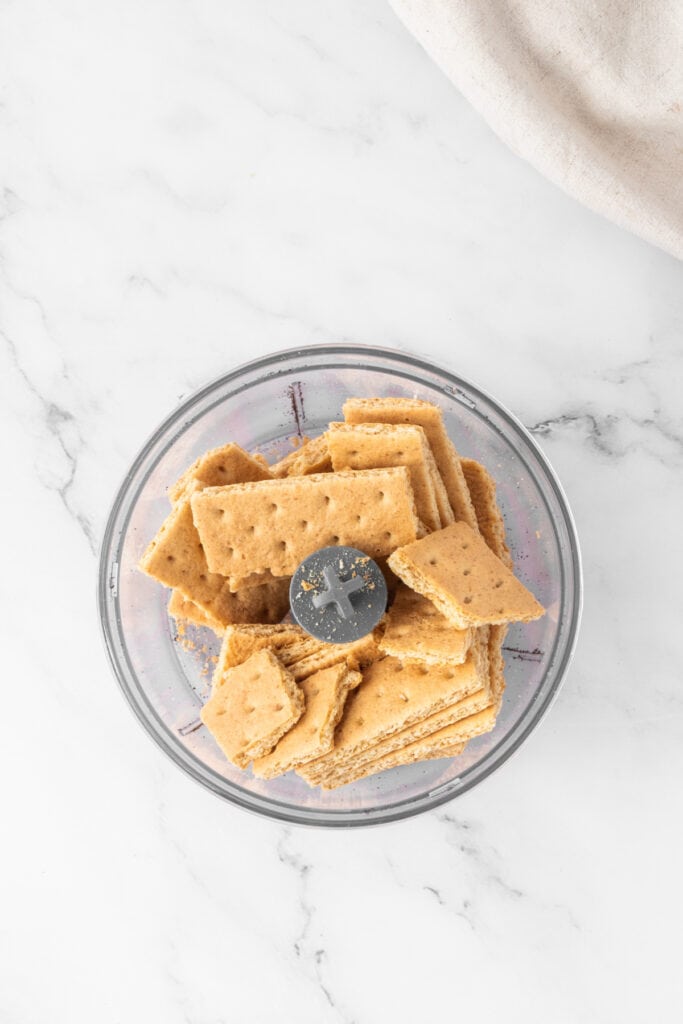 Bowl of graham crackers in a food processor ready to be pulsed for the cheesecake crust.