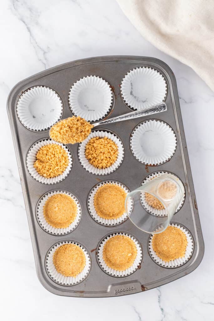 Muffin pan lined with cupcake liners filled with graham cracker crust mixture being pressed down with a small glass.