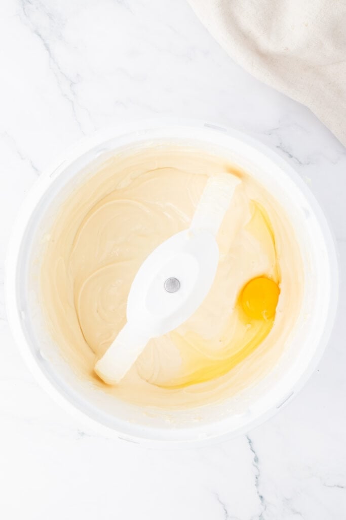 Smooth cheesecake batter in a mixing bowl with one egg being added for the next step.