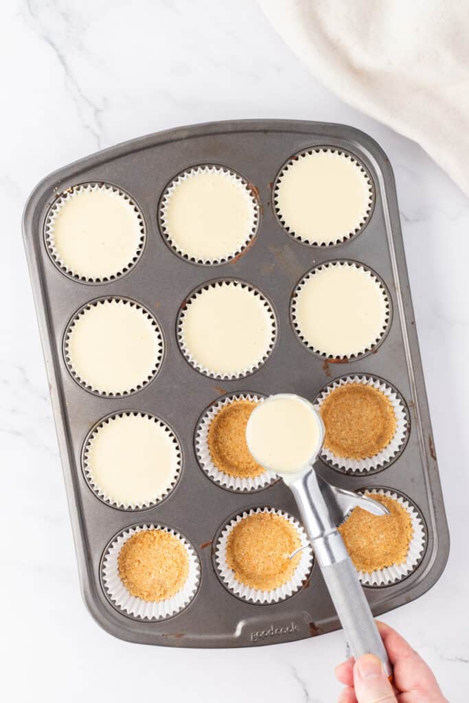 Muffin pan filled with graham cracker crusts being topped with creamy cheesecake batter using an ice cream scoop.