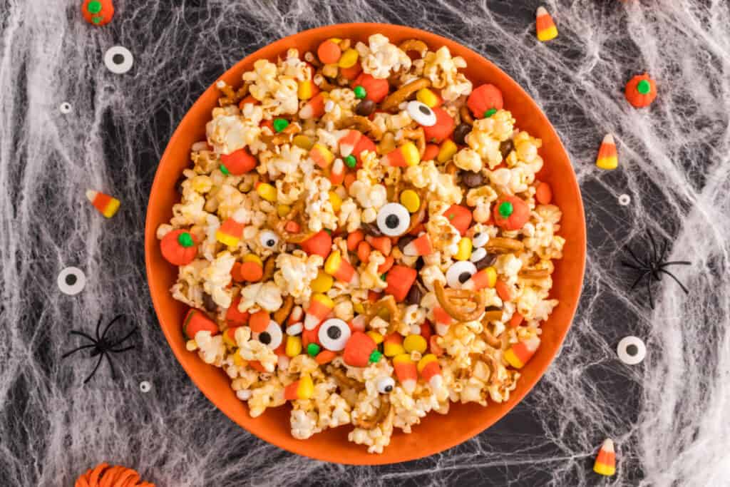 Overhead shot of Monster Mash Popcorn in an orange bowl with candy eyeballs, candy corn, and pumpkins mixed in, surrounded by Halloween decorations.