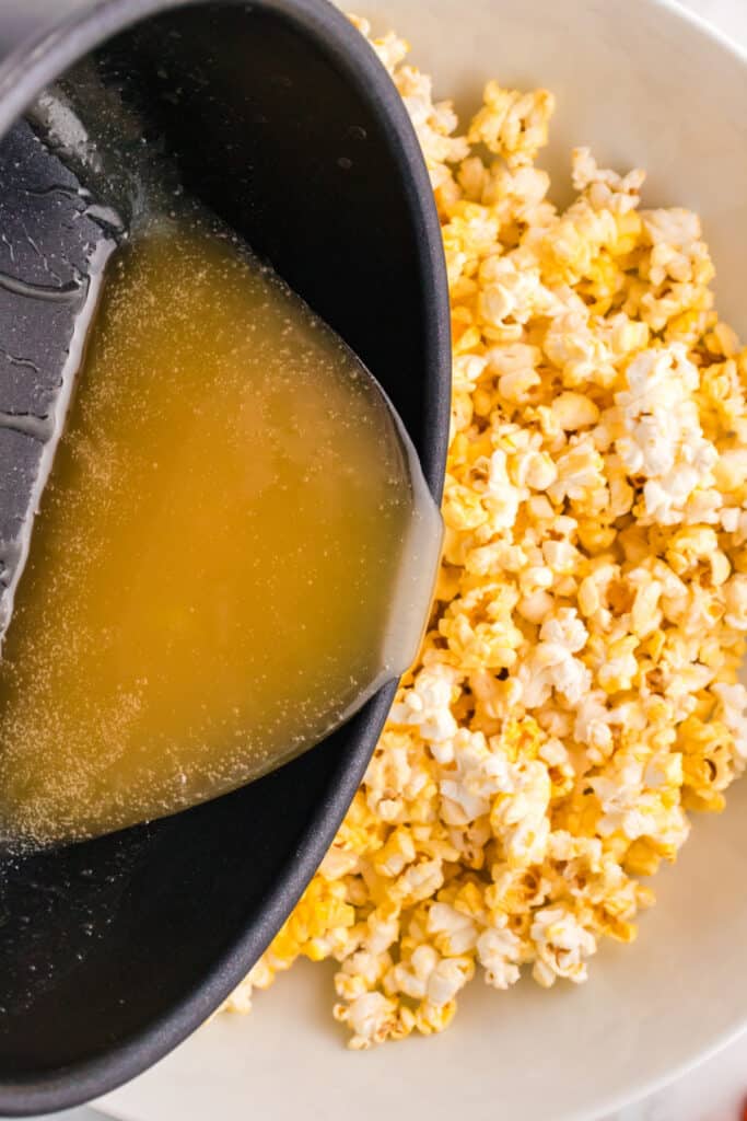 Hot syrup being poured from a saucepan over a large bowl of popcorn to coat it evenly.