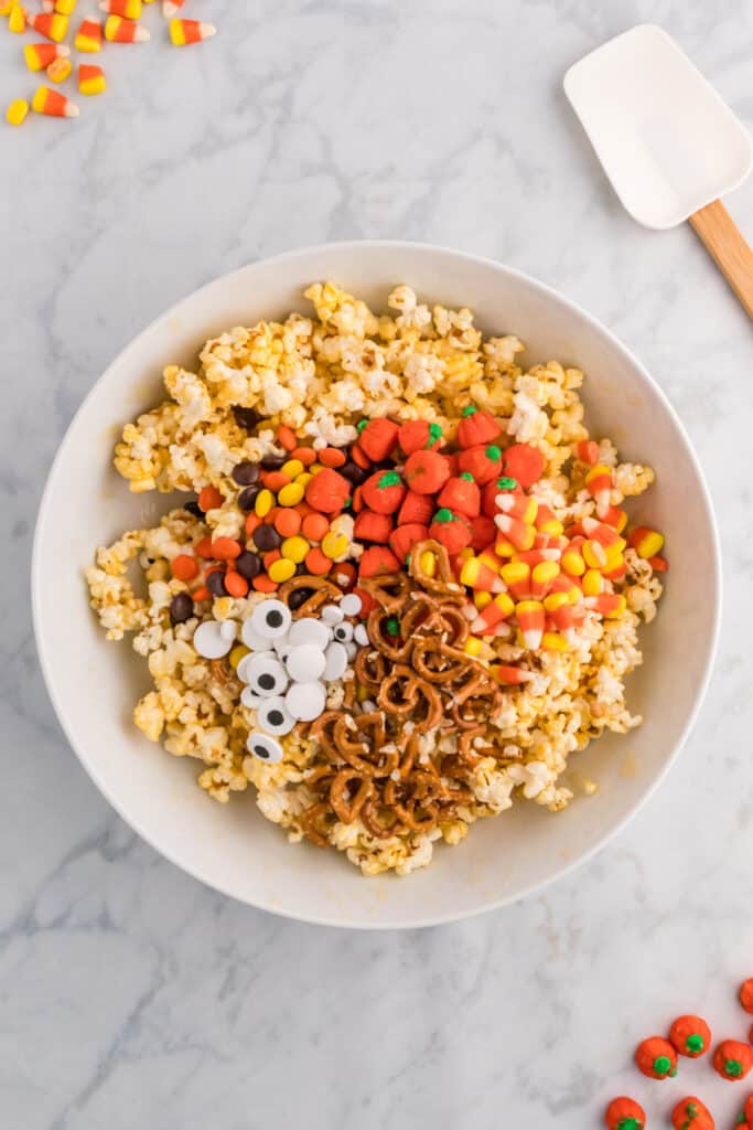 Popcorn in a mixing bowl topped with pretzels, candy corn, candy pumpkins, Reese’s pieces, and candy eyeballs before stirring.