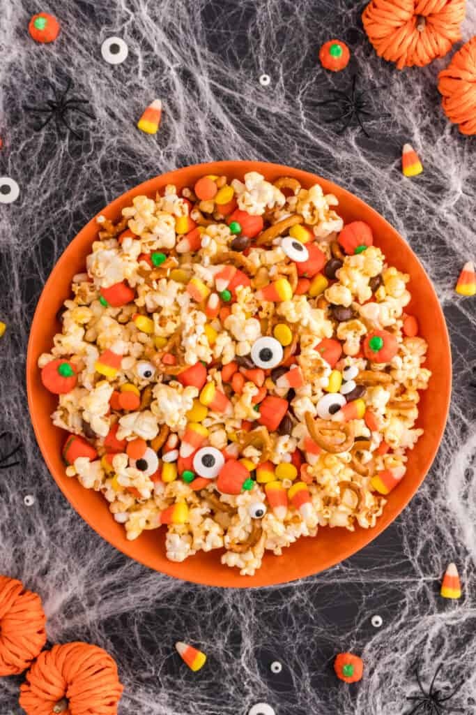 Finished Monster Mash Popcorn in an orange bowl surrounded by candy corn, candy pumpkins, and fake spiders on a spiderweb tablecloth.