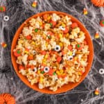 Square crop of Monster Mash Popcorn in an orange bowl with candy corn, candy pumpkins, pretzels, and eyeballs on a Halloween spiderweb background.