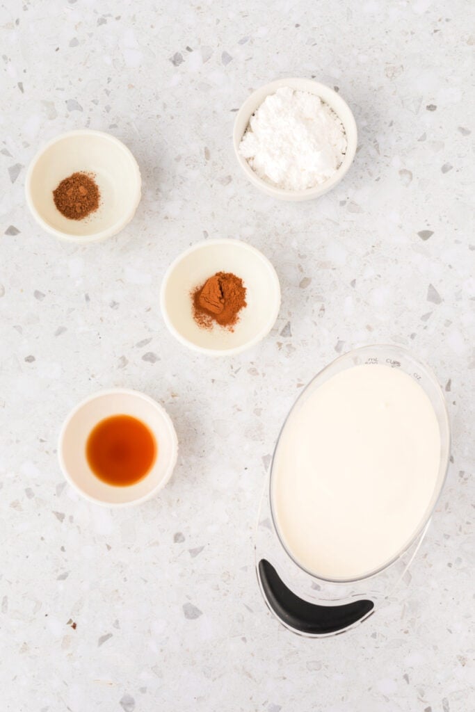 Overhead view of small bowls with ingredients for spiced whipped cream, including cinnamon, nutmeg, vanilla, powdered sugar, and cream.
