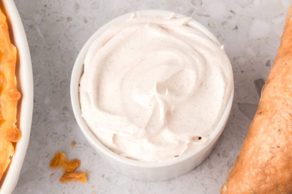 Bowl of spiced whipped cream between a baked pie and a sweet potato on a light countertop.