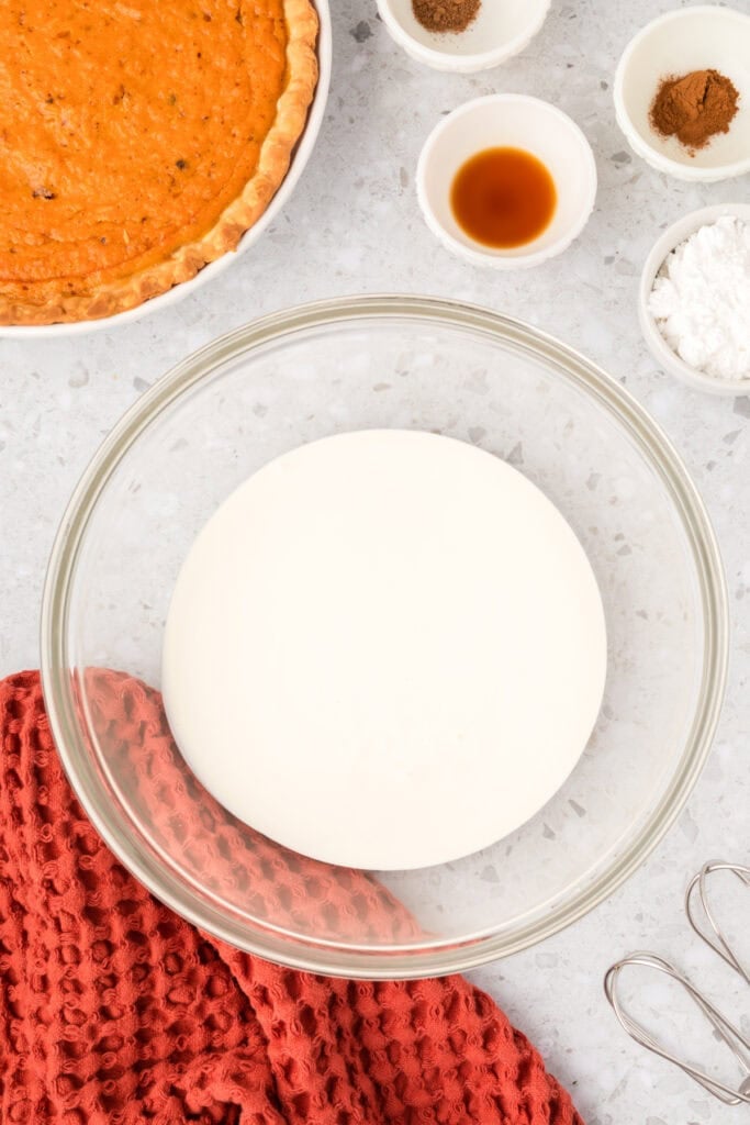 Mixing bowl filled with heavy cream surrounded by small bowls of vanilla, spices, and powdered sugar next to a sweet potato pie.