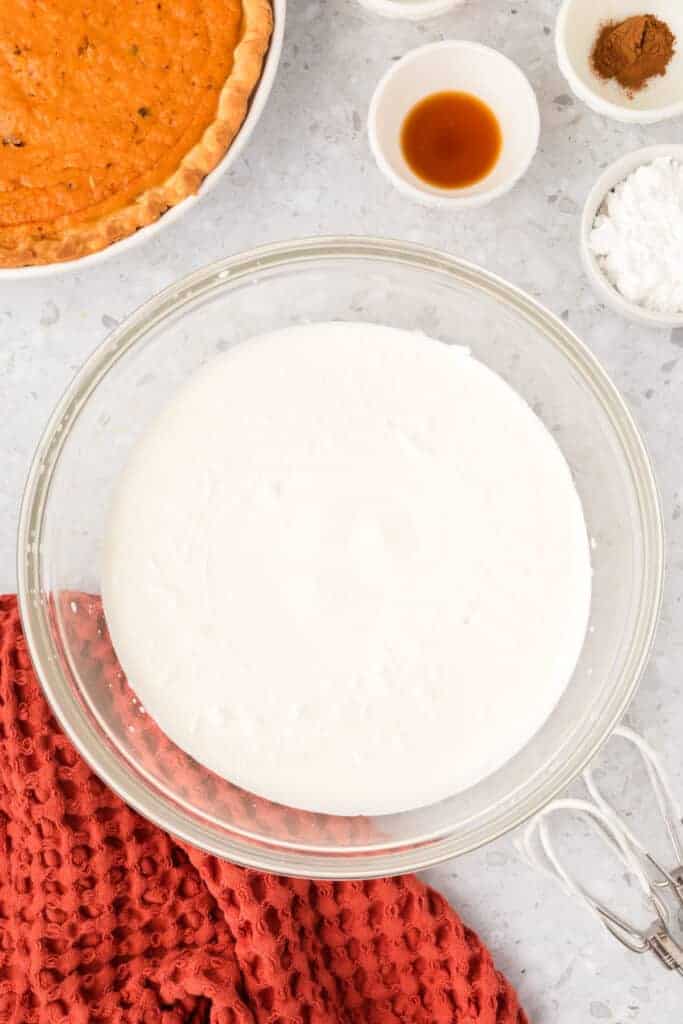 Heavy cream starting to whip in a clear bowl with ingredients and a sweet potato pie nearby.