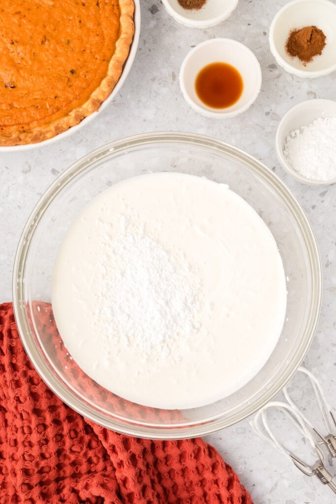 Partially whipped cream topped with powdered sugar in a glass mixing bowl beside baking ingredients.