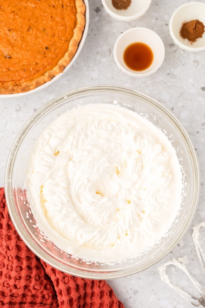 Whipped cream beaten to soft peaks in a clear bowl on a counter with vanilla and spices nearby.