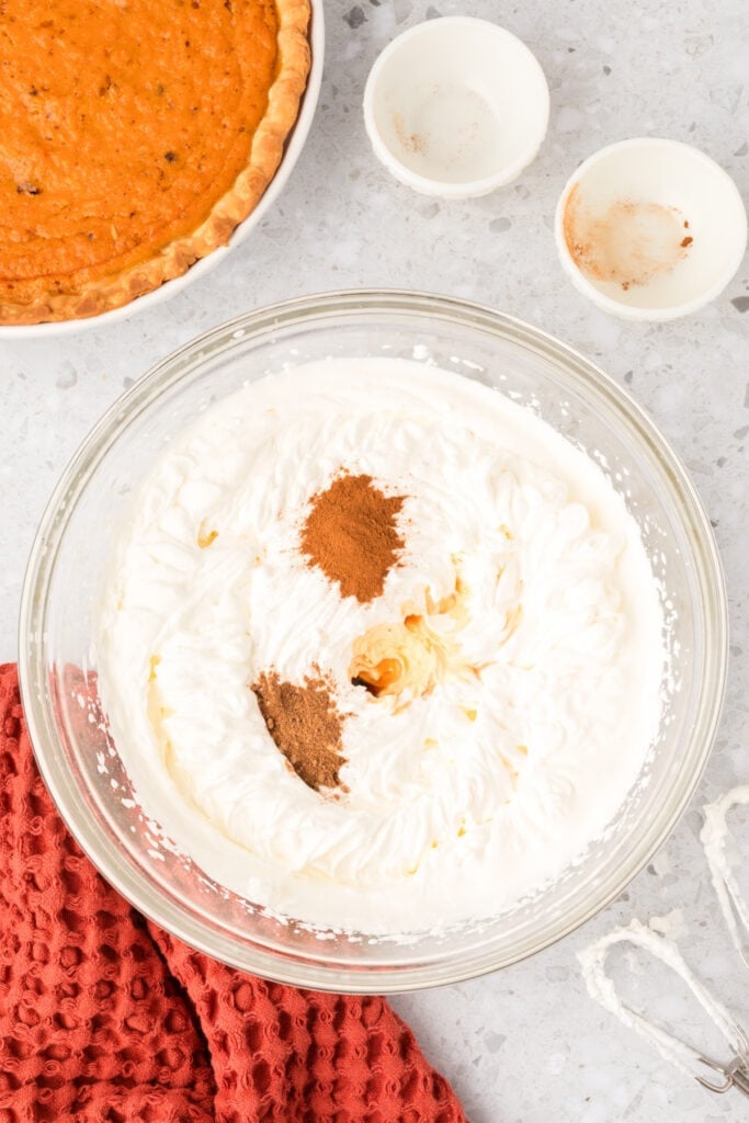 Ground cinnamon and nutmeg added to whipped cream mixture in a glass bowl ready to blend.