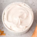 Bowl of fluffy spiced whipped cream with visible specks of cinnamon and nutmeg, sitting on a light marble surface.