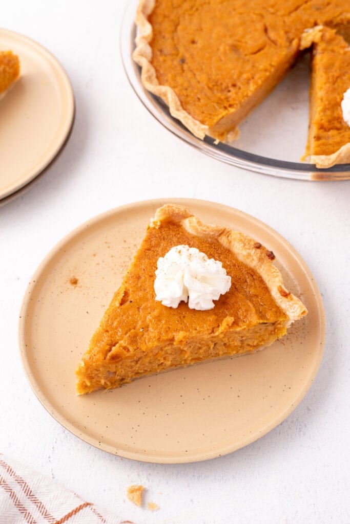 Close-up of a single slice of sweet potato pie on a tan plate topped with a swirl of whipped cream.