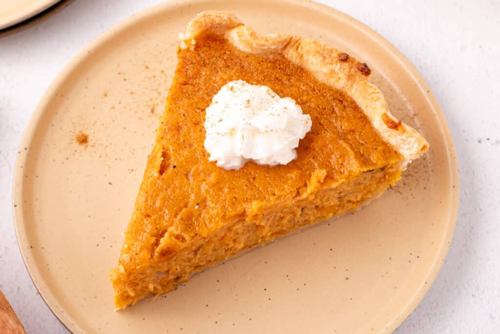 Close-up of a slice of sweet potato pie on a tan plate topped with a dollop of whipped cream and sprinkled with cinnamon.