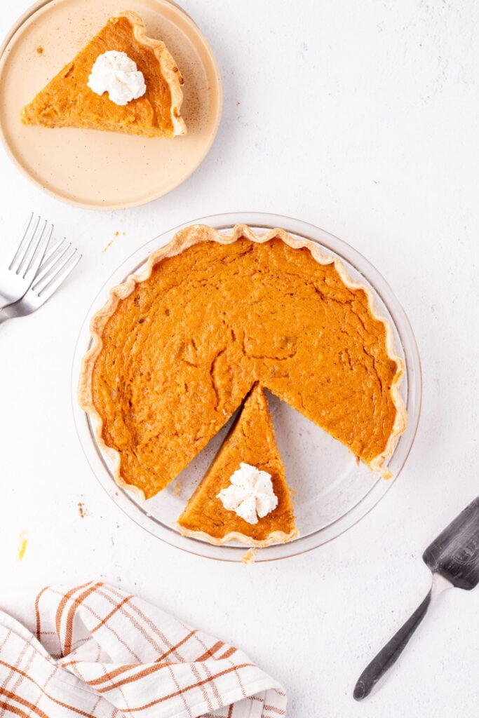 Sweet potato pie slice topped with whipped cream on a plate next to the rest of the pie.