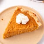 Overhead view of a slice of sweet potato pie on a tan plate with a swirl of whipped cream in the center and a flaky crust edge.
