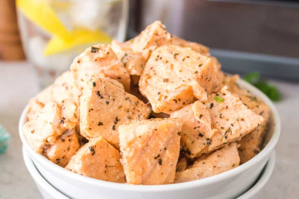 White bowl filled with seasoned, air-fried salmon bites on a countertop with an air fryer in the background.